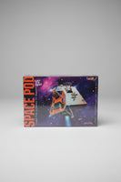 A boxed toy or game product titled 'SPACE POD' standing upright next to a black digital score counter on a light beige wall.